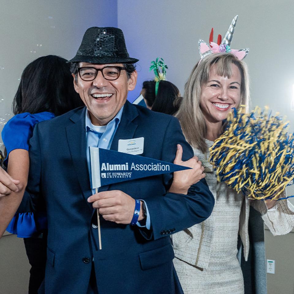 The image shows a man and a woman posing happily at an event. The man is wearing a dark suit and a black sequined hat, holding a small flag that reads "Alumni Association St. Edward's University." The woman is smiling and holding blue and yellow pom-poms, wearing a unicorn headband. Both appear to be enjoying themselves in a festive atmosphere, with bubbles and decorations in the background, suggesting a fun and celebratory gathering.