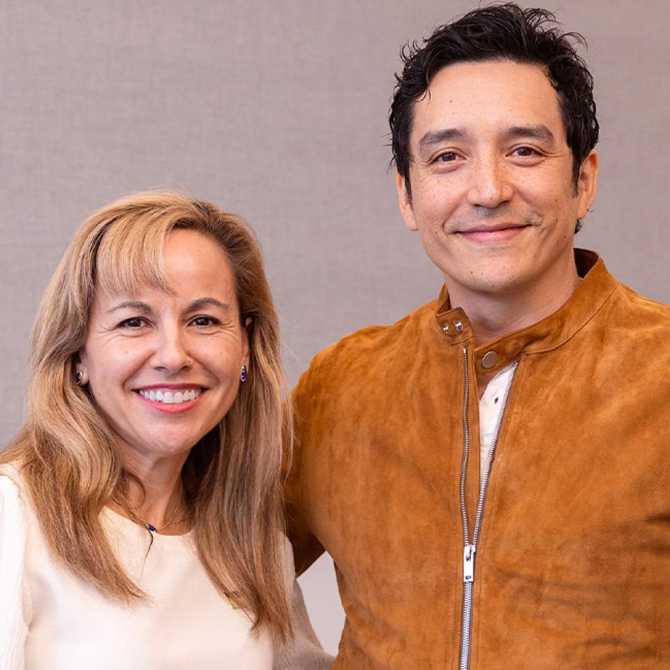 The image shows two people posing for a photo. On the left is a woman with blonde hair wearing a white top, and on the right is a man with dark hair wearing a brown suede jacket. They are both smiling at the camera. The setting appears to be indoors, with a neutral background that includes a light-colored wall and possibly a piece of artwork. The atmosphere is friendly and informal, suggesting a casual meeting or visit.