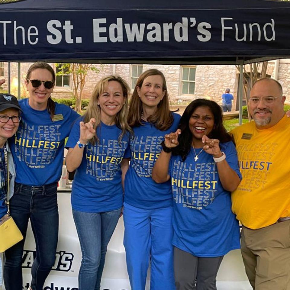 The image features six people standing together under a canopy that reads "The St. Edward's Fund." They are all smiling and making hand gestures, likely the university's hand sign. Most of them are wearing blue T-shirts with "HILLFEST" printed on them, indicating they are part of the same event. One person is wearing a yellow version of the T-shirt. The background shows a campus setting with buildings and greenery, suggesting this is a university event.