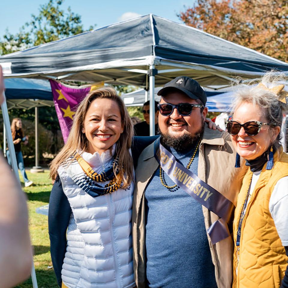 The image shows three people posing for a photo at an outdoor event. The person in the middle is wearing a sash that reads "University" and sunglasses, while the two women on either side are smiling and dressed in casual outdoor clothing, with the woman on the left wearing a white vest and the one on the right in a yellow vest. The background features several tents and people, suggesting a festive, social gathering such as a homecoming event. A fourth person is taking their picture with a smartphone.