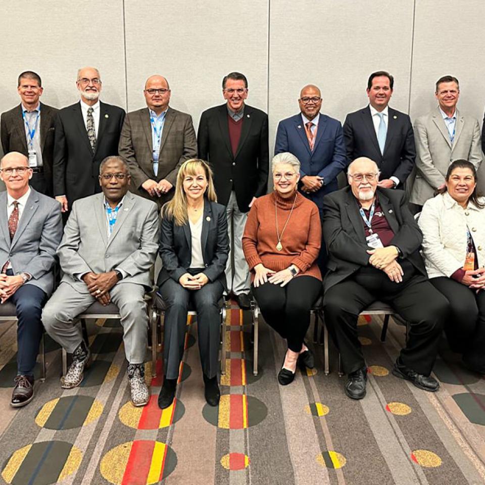 The image shows a group of seventeen individuals posing for a formal group photo at a conference. They are arranged in two rows, with the front row seated and the back row standing. Everyone is dressed in business attire, including suits and blazers. The background is a plain conference room wall, and the carpet has a colorful, modern design. The individuals are smiling, suggesting a positive and professional atmosphere, likely related to a meeting or conference event.