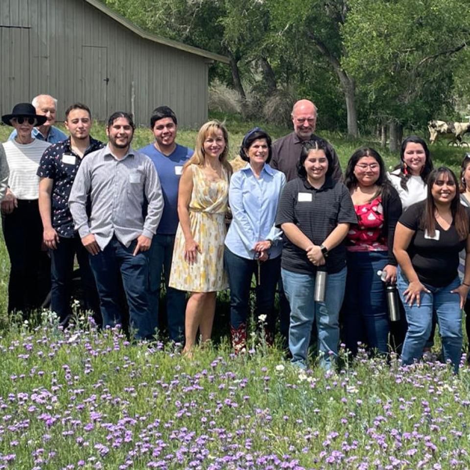 The image shows a group of people standing outdoors in front of a wooden building, likely on a ranch. They are posing for a group photo, standing on a field of purple wildflowers. The group consists of men and women, dressed casually, suggesting a relaxed, possibly educational or team-building event. The background includes trees and a cow, reinforcing the rural setting. The atmosphere appears friendly and informal, with everyone smiling for the camera.