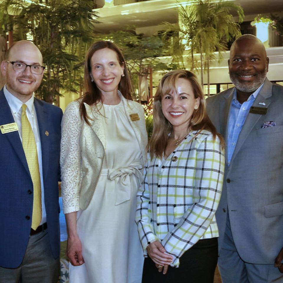 The image shows four individuals standing together outdoors in a well-lit, lush garden setting. They are dressed in business attire, including suits and a dress. The group consists of three men and one woman, all smiling at the camera. The woman is wearing a plaid blazer, while the men are in suits with ties. The setting appears to be a formal event or gathering, possibly related to a conference or professional meeting. The atmosphere is friendly and professional.