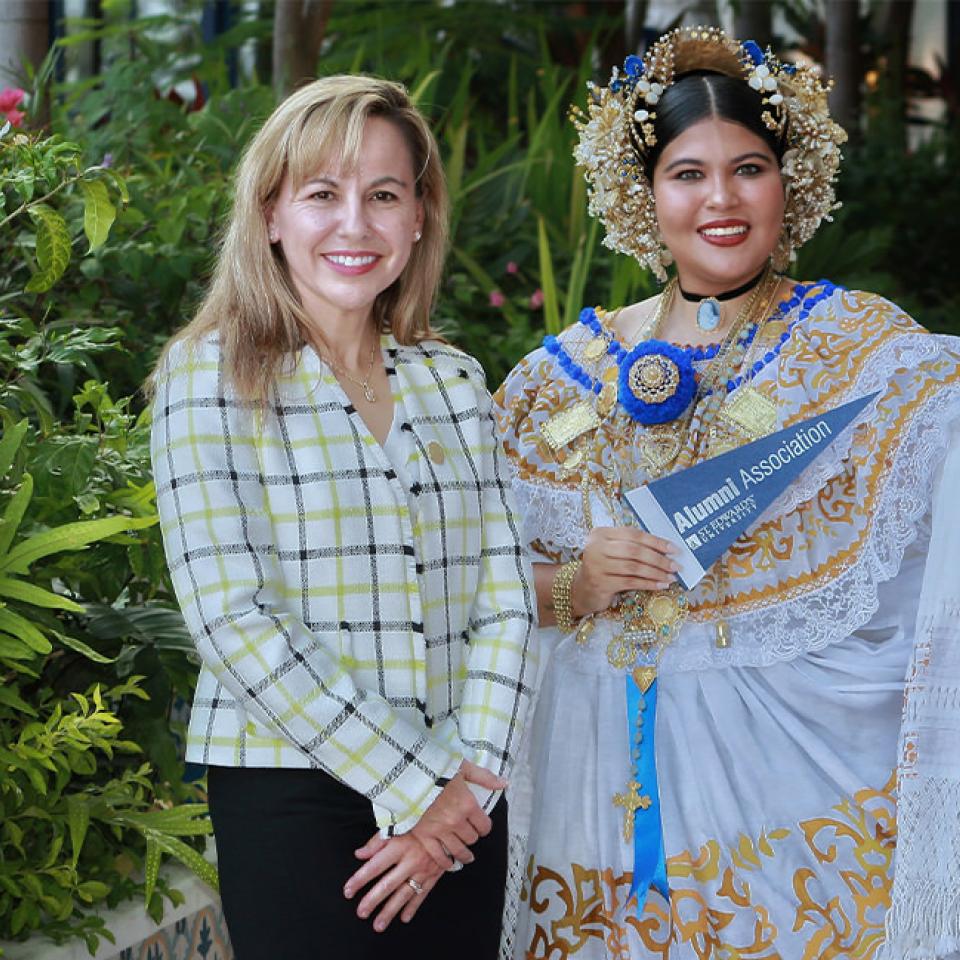 The image shows two women standing outdoors in front of lush greenery. The woman on the left is dressed in business attire, wearing a plaid blazer and black skirt. The woman on the right is dressed in an elaborate traditional Panamanian dress with gold and blue embroidery and ornate headpieces. She holds a sign that reads "Alumni Association." Both women are smiling at the camera, suggesting a celebratory or cultural event, possibly related to alumni activities or a cultural showcase.