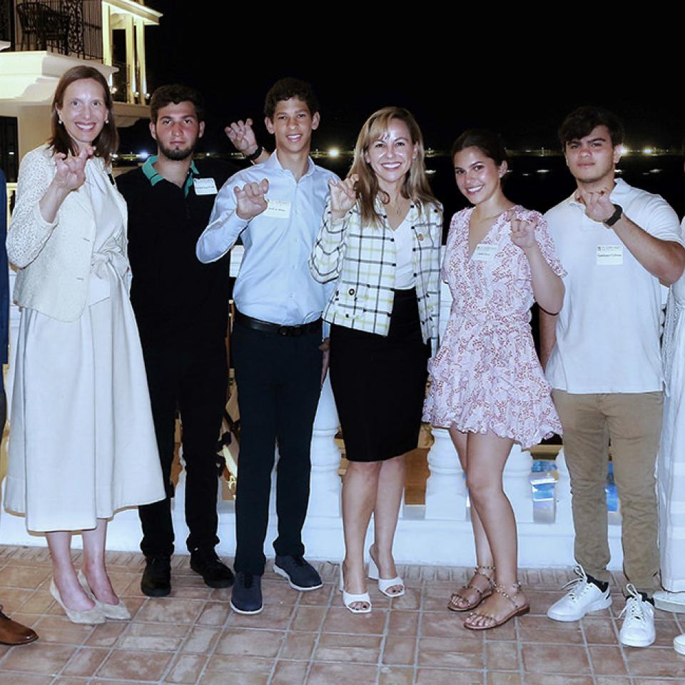 The image shows a group of eight people standing on a balcony at night. They are smiling at the camera. The group includes both adults and young adults, dressed in a mix of business casual and casual attire. The background features a well-lit building and a view of lights in the distance, suggesting a scenic or waterfront location. The setting and gestures indicate a celebratory or school-related event, likely involving St. Edward's University.