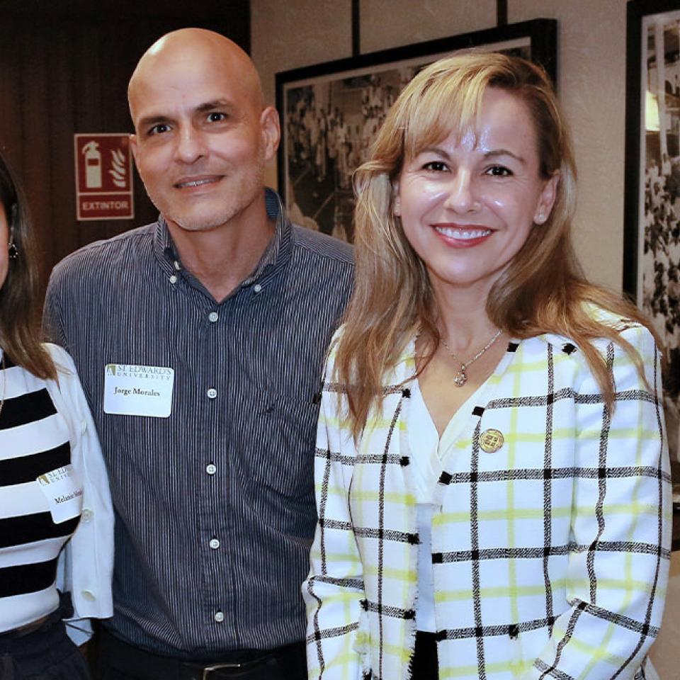 The image shows three people smiling at the camera indoors. The woman on the left is wearing a black and white striped top, the man in the middle is in a blue striped shirt, and the woman on the right is wearing a plaid blazer. They are standing closely together, and the man and woman on the right are wearing name tags, indicating a formal or semi-formal event, likely related to St. Edward's University as suggested by their name tags. The background features framed black-and-white photos.