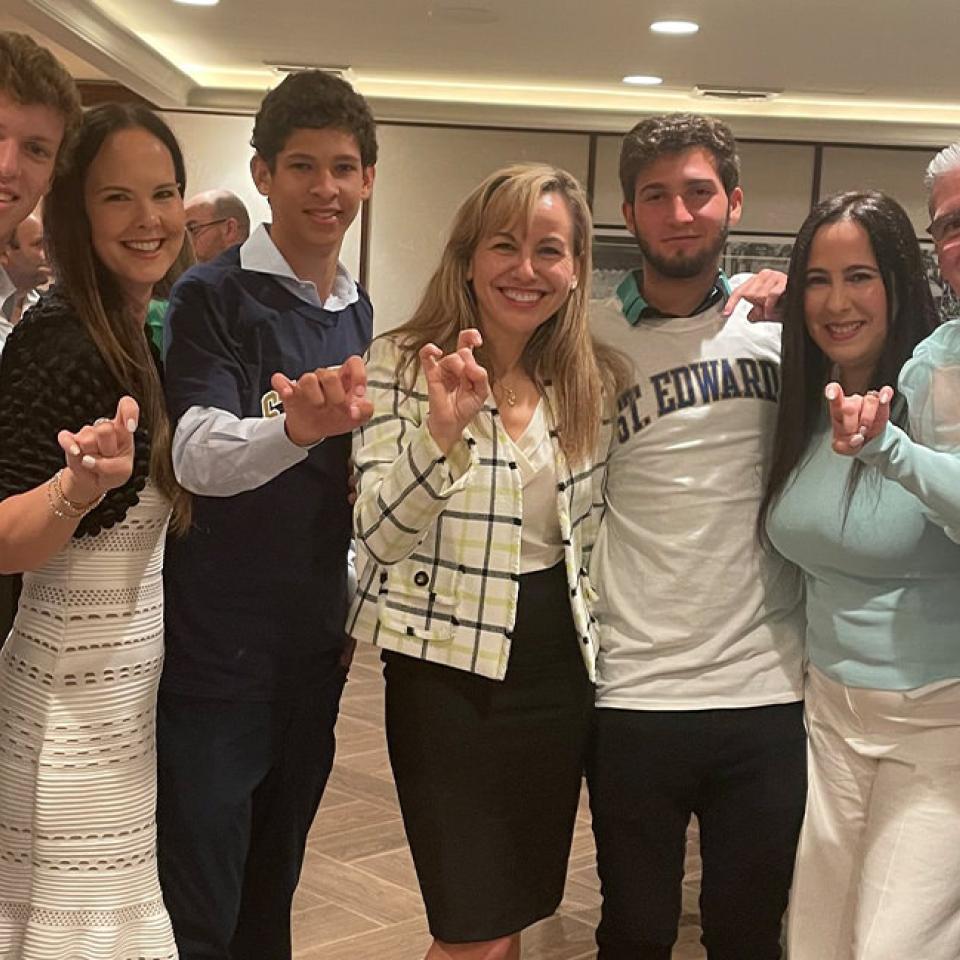 The image shows seven people standing closely together indoors, smiling at the camera, and making the "hook 'em horns" hand gesture. They are dressed in a mix of casual and business casual attire. The group includes both young adults and older individuals, with one person wearing a "St. Edward's" shirt, suggesting they might be students or alumni of St. Edward's University. The setting appears to be a social or celebratory event, possibly related to the university.