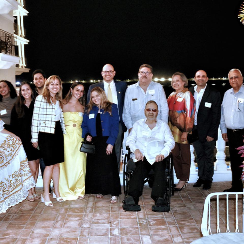 The image shows a group of people posing together on a balcony at night. They are dressed in a mix of formal and semi-formal attire. Among them is a woman in a traditional Panamanian dress, indicating a cultural event or celebration. The group includes men and women of various ages, some in suits and others in dresses. The background shows a building and a view of lights in the distance, suggesting a scenic or waterfront location. The atmosphere is festive and celebratory.