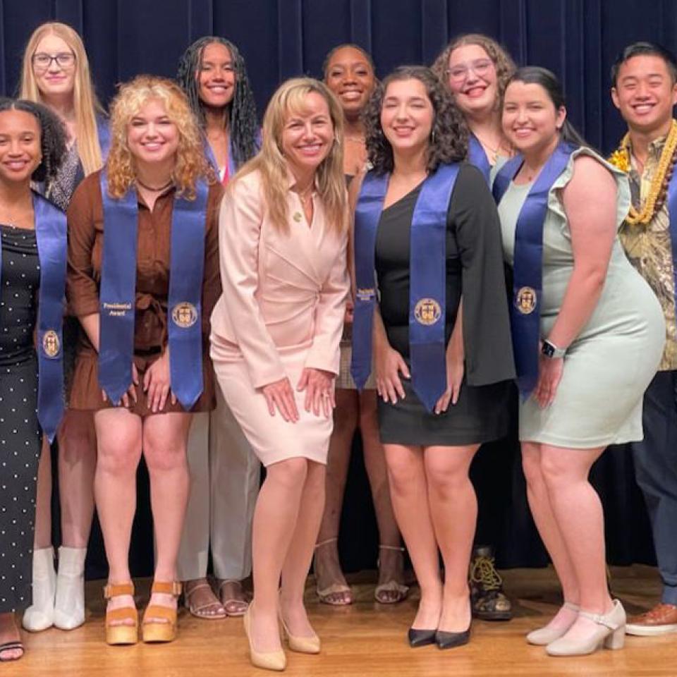 The image features a group of eleven people posing together on a stage. They are dressed in formal attire, with several individuals wearing blue sashes that read "Presidential Award." In the center of the group is a woman in a light pink suit, who appears to be the focus of the photo. The group is smiling, suggesting a celebratory occasion, likely an awards ceremony or similar event. The backdrop is a dark curtain, adding a formal touch to the setting.