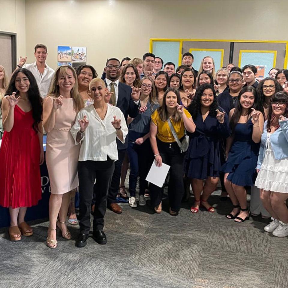 The image shows a large group of people, mostly young adults, posing together in a room. They are dressed in a mix of formal and semi-formal attire, with many making a hand gesture, likely a university symbol. The group includes both men and women, and they are all smiling, suggesting a celebratory occasion. The setting appears to be an indoor event, possibly a ceremony or reception, as indicated by the formal clothing and the overall festive atmosphere.