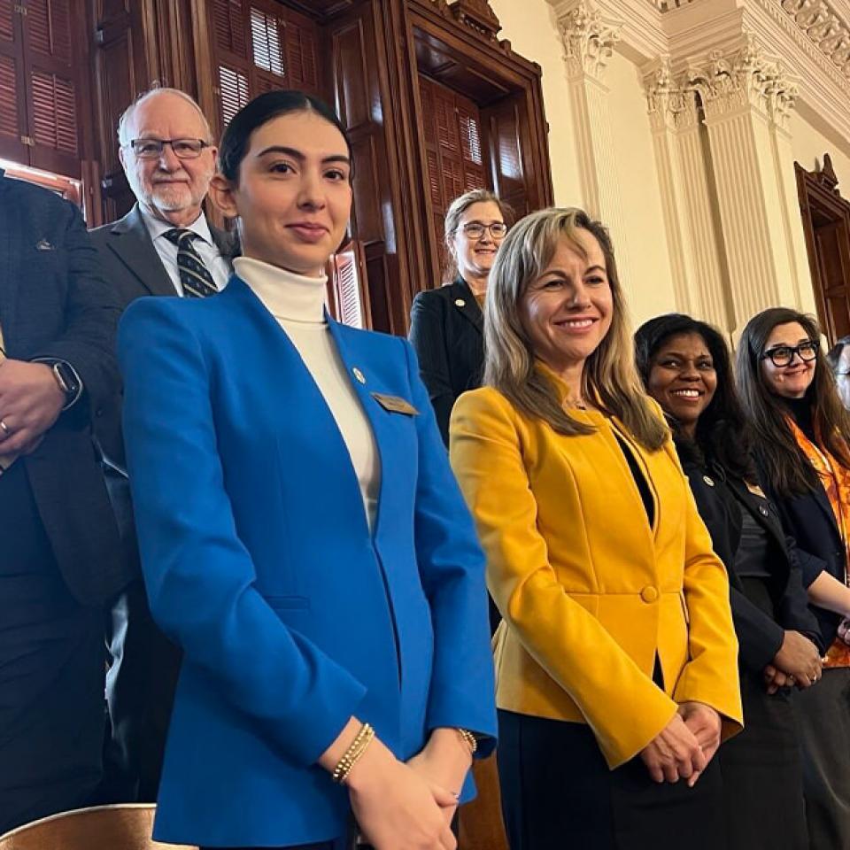 The image shows a group of eight people standing in what appears to be a formal setting, likely a government building, given the ornate interior and wooden fixtures. They are positioned on steps and smiling at the camera. The individuals are dressed in professional attire, with the two women in the foreground wearing bright blazers, one in blue and the other in yellow. The setting and their attire suggest they are participating in a professional event or a visit to the state capitol.