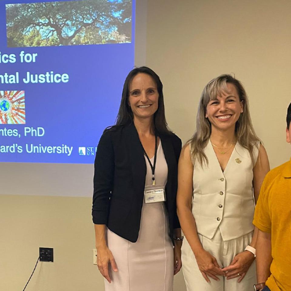 The image shows three people standing in front of a projected presentation slide titled "Statistics for Environmental Justice" by Montse Fuentes, PhD, President of St. Edward's University. The woman on the left is wearing a black blazer and light dress, Montse Fuentes in the middle is wearing a white sleeveless suit, and the man on the right is wearing a yellow polo shirt. They are all smiling and posing for the photo, indicating they are likely attending or participating in a conference or seminar.