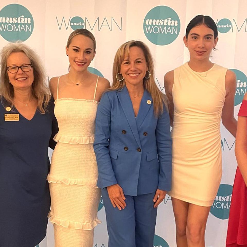 The image shows six women standing in front of a backdrop with the logos "austin WOMAN" and "WOMAN." They are all dressed in elegant attire, including dresses and a suit. From left to right, the first woman is wearing a green dress, the second a dark blue dress, the third a white dress, the fourth a blue suit, the fifth a white dress, and the sixth a red dress. They are all smiling and appear to be at a formal event, possibly an awards ceremony or gala.