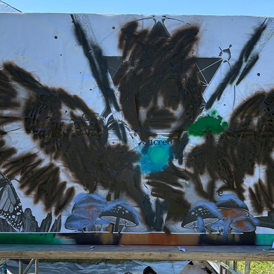 The image depicts a large mural in progress, featuring the outline of a person with wings and an owl headpiece. The figure's details are faintly visible, with sections filled in with dark brown paint. Surrounding elements include butterflies and mushrooms. The mural is set up outdoors on a grassy area, with a blue canopy tent in the background and art supplies scattered nearby. The scene captures the early stages of the mural's creation.