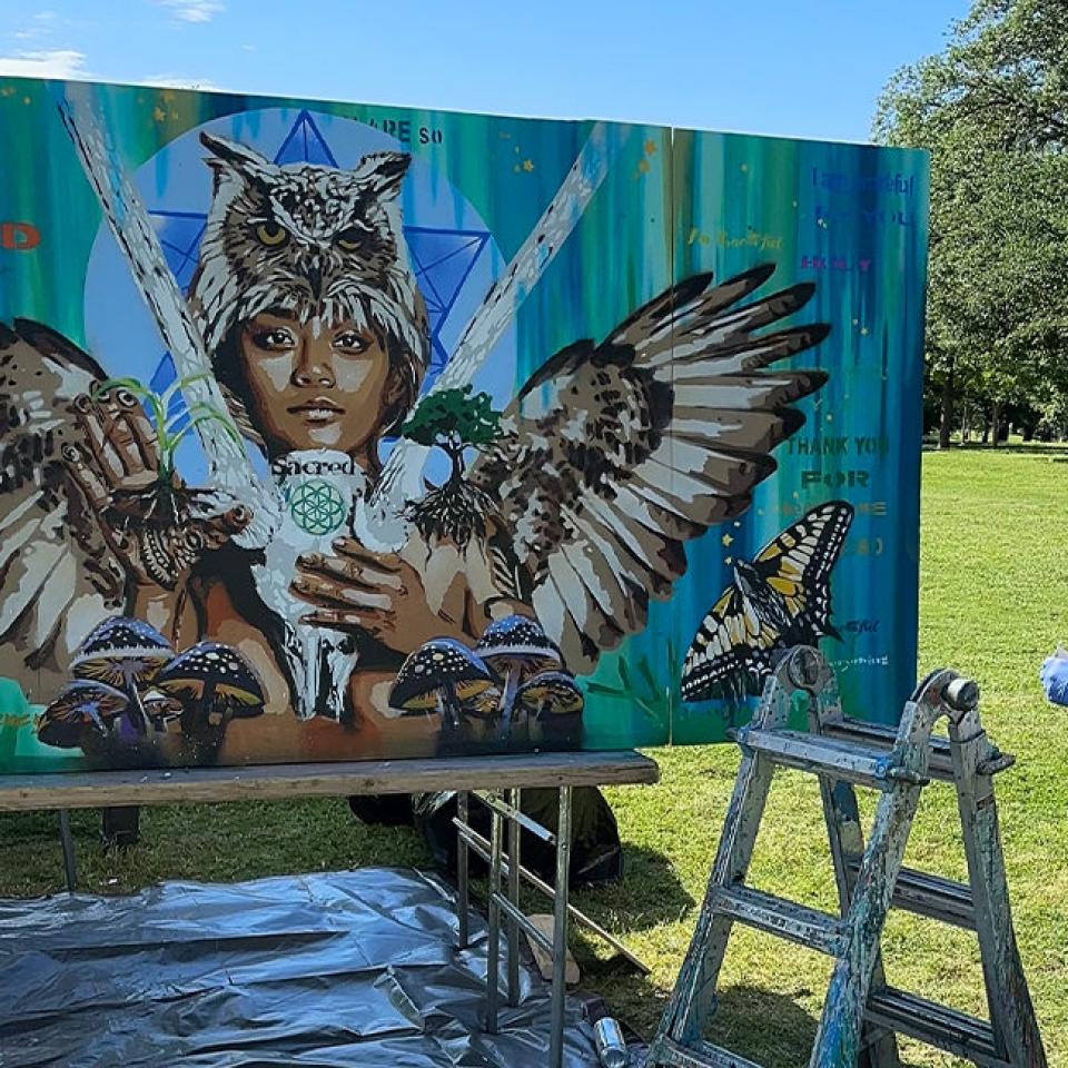 The image shows an artist standing outdoors in a park, working on a large mural. The mural features a central figure with an owl on their head, wings spread wide, and holding various nature elements like plants and mushrooms. The background includes butterflies and words such as "kind," "thank you," and "sacred." The artist, wearing a camouflage cap and black tank top, is standing next to a ladder and appears to be making finishing touches. The setting is bright and sunny, indicating a live art event.