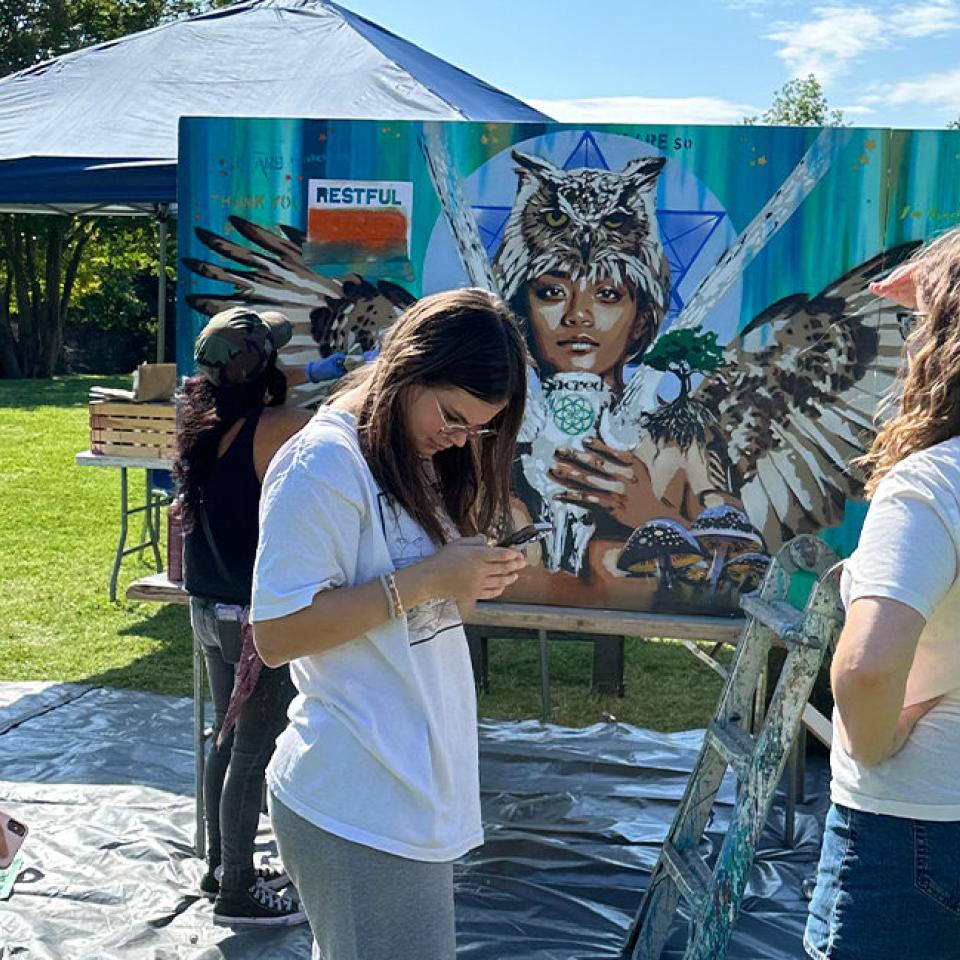 The image shows an outdoor art session where a muralist is actively painting a large canvas depicting a person with wings and an owl headpiece. The muralist, wearing a cap and working intently, is accompanied by three onlookers. Two of them, dressed casually, are observing the artwork, while one is engrossed in their phone. The setup includes a blue canopy tent, art supplies, and a grassy park setting with trees in the background.