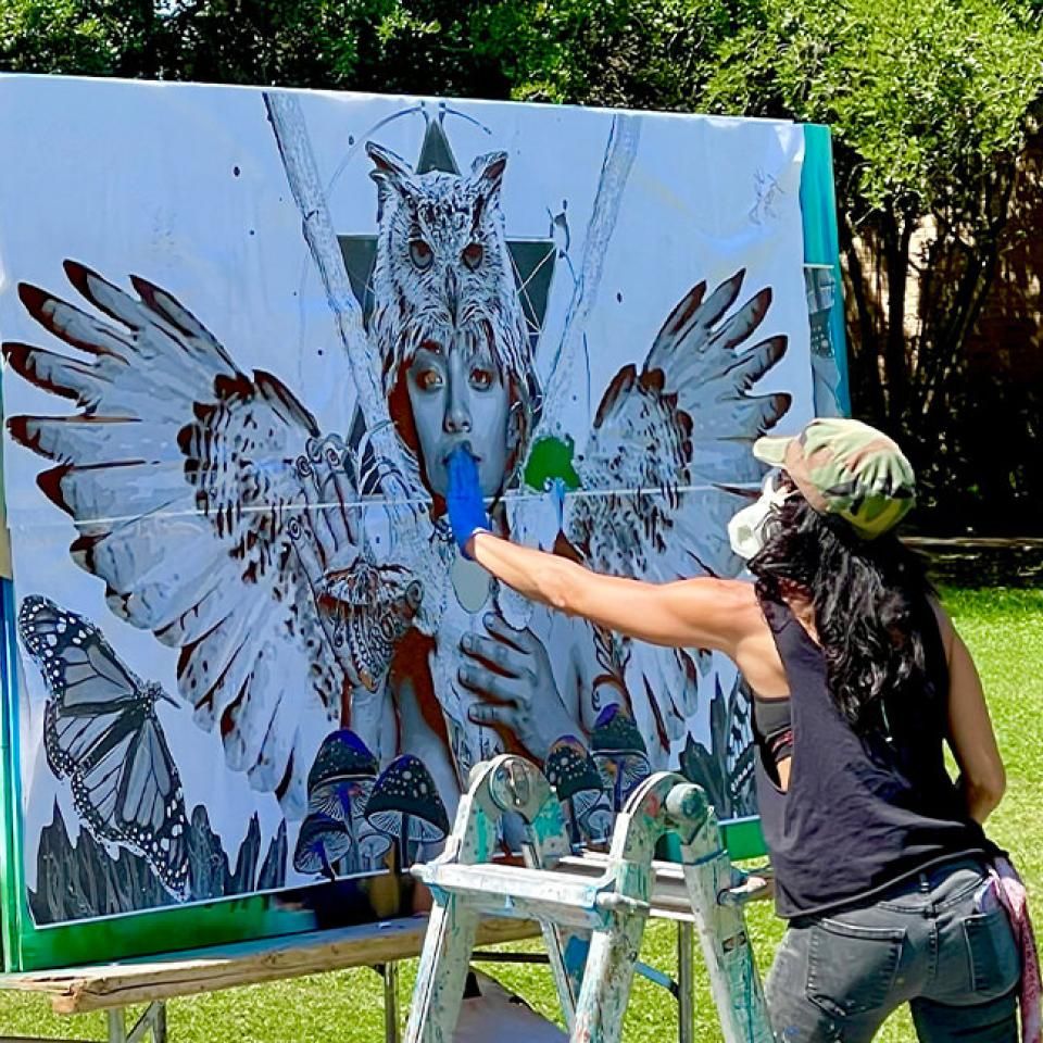 The image shows an artist working on a large mural outdoors in a park. The mural depicts a central figure with an owl on their head and wings spread wide, holding nature elements like plants and mushrooms. The artist, wearing a camouflage cap and black tank top, is using a spray can to add details to the mural. She is standing on a ladder, wearing a face mask for protection. The setting is sunny with trees in the background, indicating a live art event or festival.