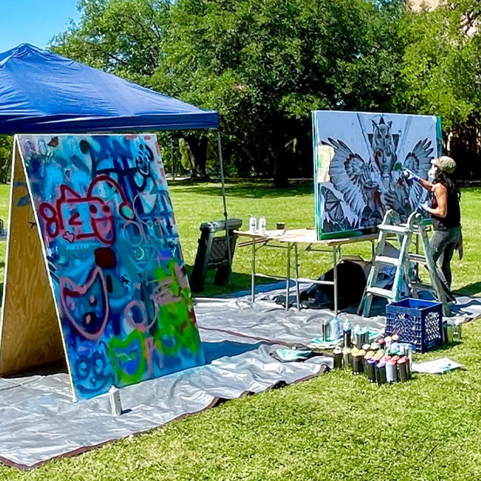 The image depicts an outdoor art setup where a muralist is actively working on a large canvas. Under a blue canopy tent, there are two canvases: one featuring colorful graffiti art with playful shapes and another showcasing a detailed mural of a person with wings and an owl headpiece. The artist is standing on a ladder, painting the mural. Various spray paint cans and art supplies are scattered around the area, and the scene is set on a grassy field with trees in the background.