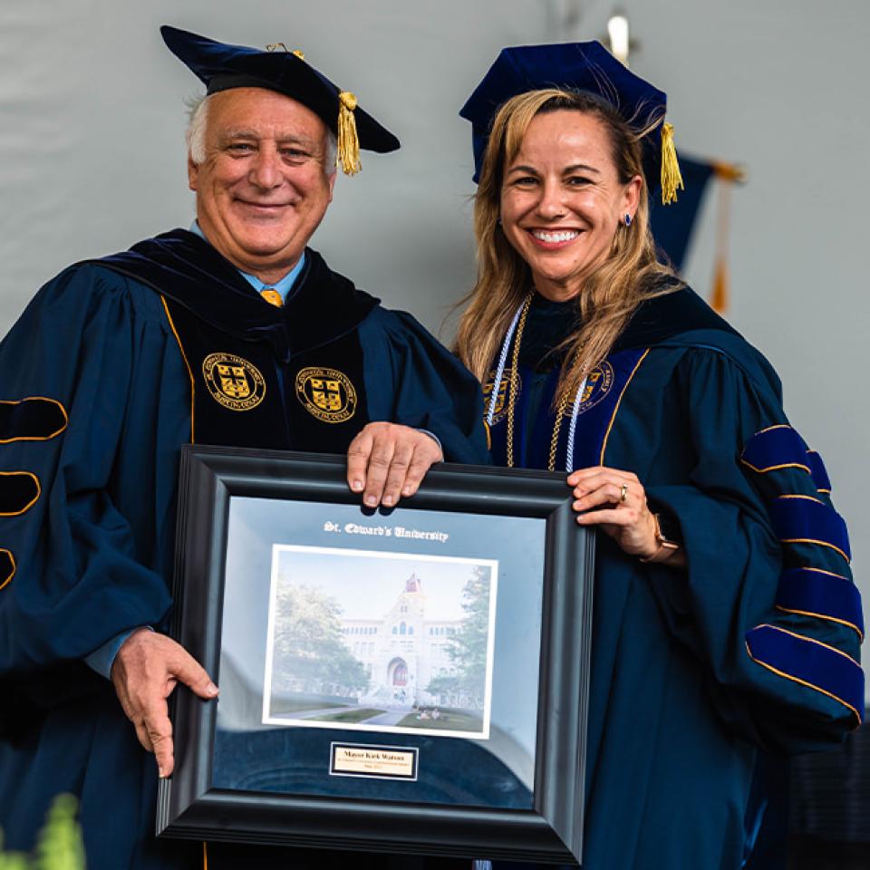 The image shows two individuals dressed in academic regalia, smiling and posing for a photo during a formal event. The man on the left is holding a framed picture of a building labeled "St. Edward's University." Both are wearing dark blue gowns with gold accents and tassels on their caps, indicating a commencement or graduation ceremony. The setting appears to be on a stage with a decorative backdrop and other attendees in similar attire in the background.