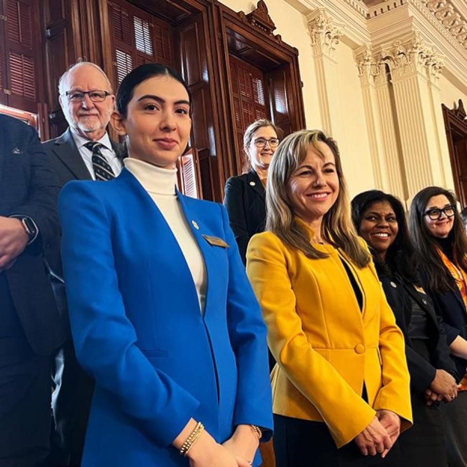 The image shows a group of eight people standing inside a government building, likely a legislative chamber. They are posing for a group photo and smiling. The group includes five women and three men, all dressed in professional attire. Notably, one woman is wearing a bright blue blazer, another a yellow blazer, and the rest are in various business suits. The background features ornate woodwork and tall windows.