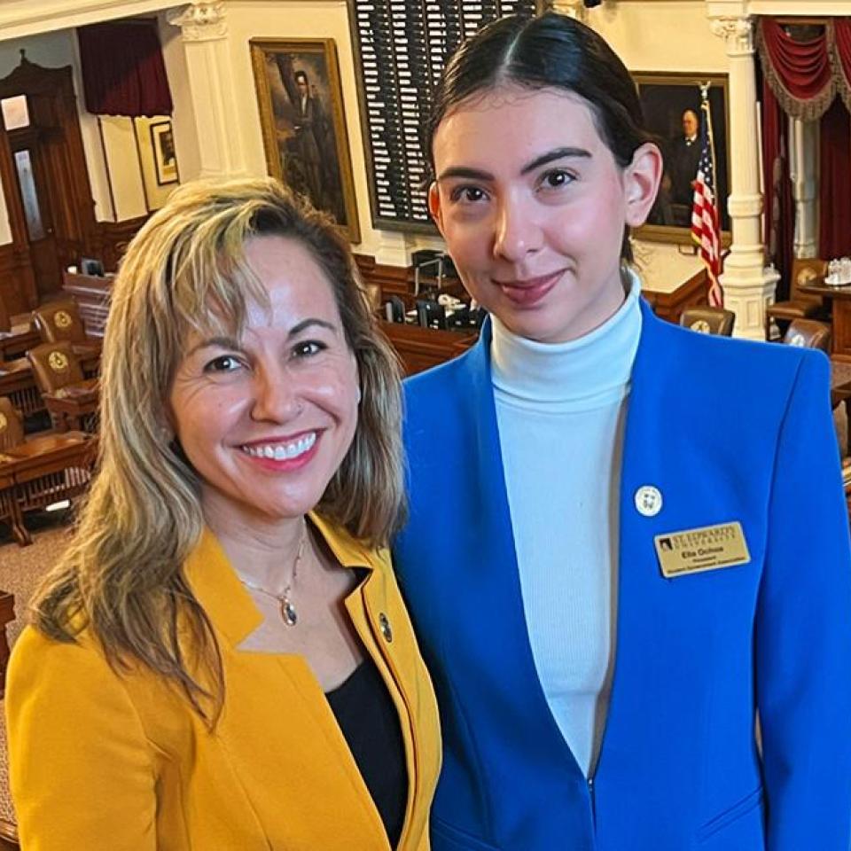 The image shows two women standing together in a legislative chamber. They are smiling and dressed in professional attire. One woman is wearing a bright yellow blazer, and the other is wearing a bright blue blazer with a white turtleneck. The background features wooden desks and chairs, a large display board, and American flags. The setting appears to be an official government building.