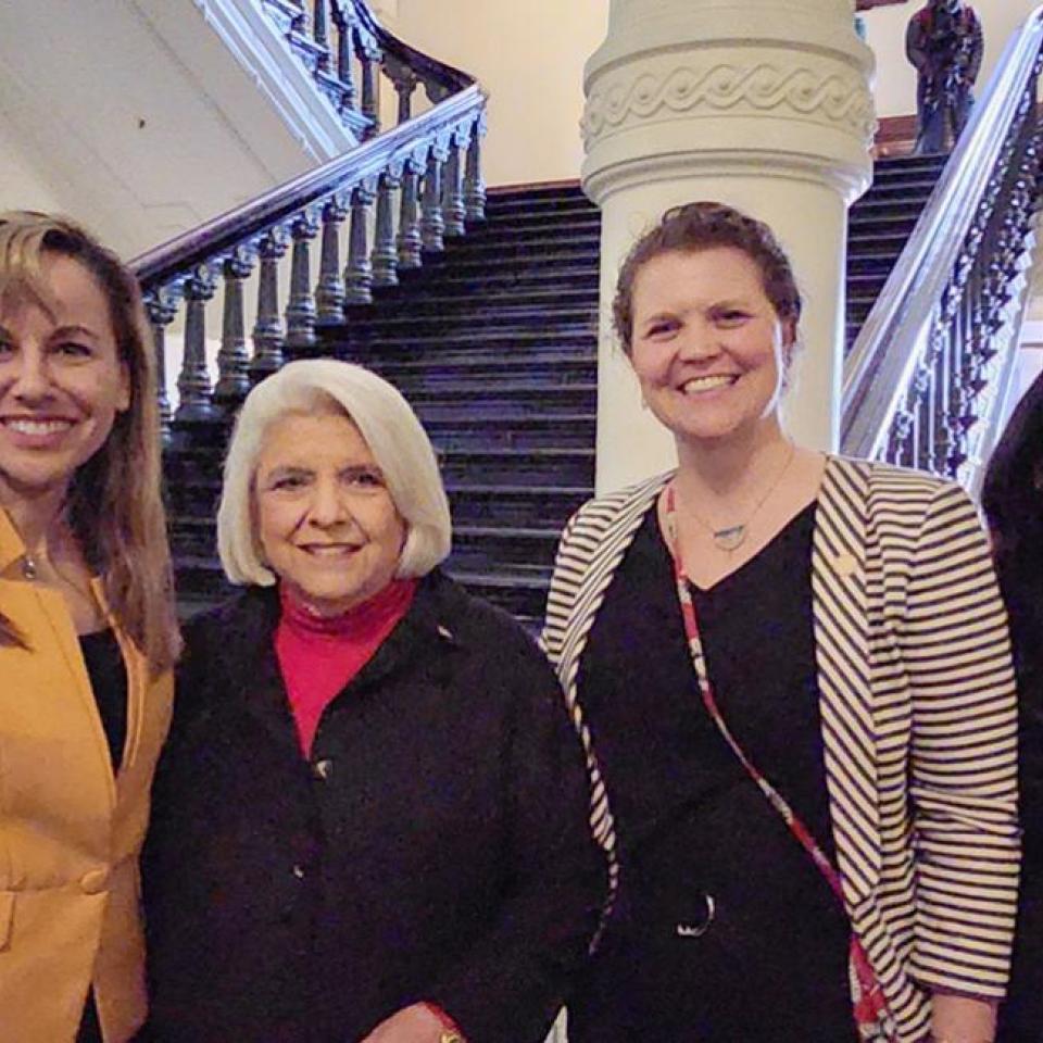 The image shows four women standing together in front of a grand staircase inside a government building. They are smiling and dressed in professional attire. One woman is wearing a yellow blazer, another is in a black outfit with a red shirt, the third is in a black dress with a striped jacket, and the fourth is in a black blazer. The background features ornate architectural details, including columns and a staircase with intricate railing.