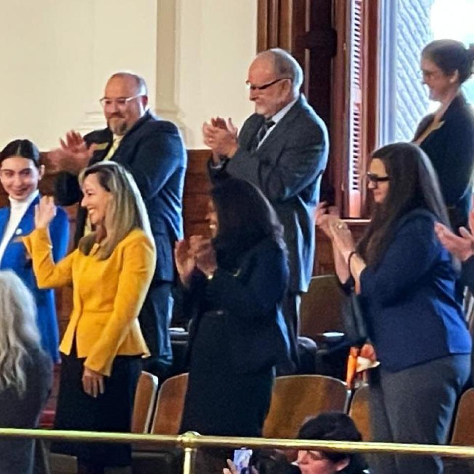 The image shows a group of people standing and clapping in a legislative chamber. They are dressed in professional attire, and some are smiling while applauding. Among them, two women are prominently visible: one wearing a bright blue blazer and another in a bright yellow blazer. The atmosphere appears to be celebratory or in recognition of an event or person, with the group standing in the seating area of the chamber.