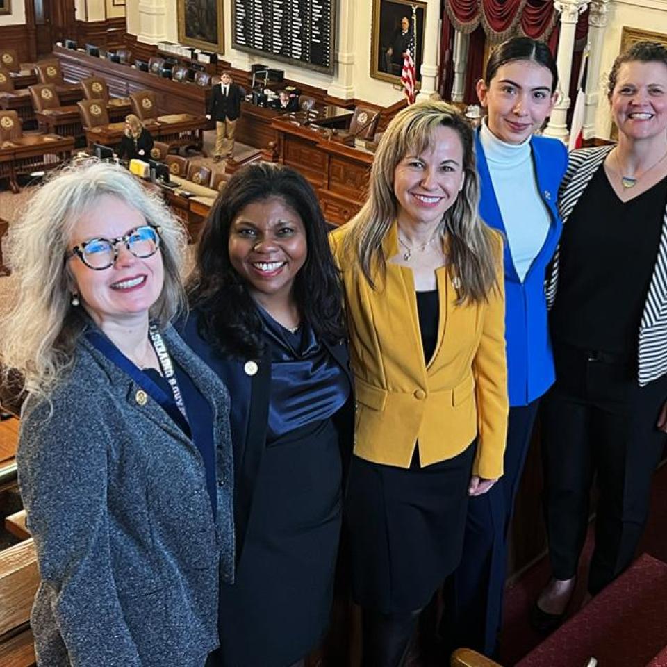 The image shows five women standing together in a legislative chamber, likely a government building. They are smiling and appear to be posing for a group photo. The chamber has rows of wooden desks and chairs, a large display board on the wall, and American flags in the background. The women are dressed professionally, with one wearing a yellow blazer, another in a blue blazer, and the others in dark attire.