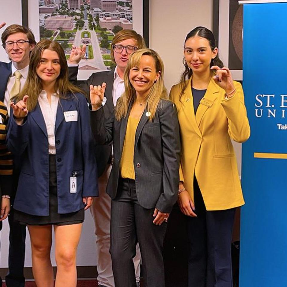 The image shows six individuals standing together in front of a banner for St. Edward's University. They are posing for a group photo, with some making a hand gesture. The group includes three women and three men, all dressed in professional or semi-professional attire. One woman is wearing a yellow blazer, and another is in a dark suit. The background includes a large photo of a university campus.