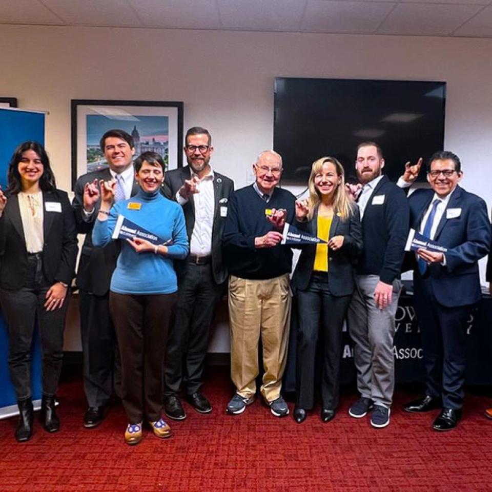 The image shows a group of eleven people standing together in a room, posing for a group photo. They are making a hand gesture and smiling. The group includes men and women, all dressed in professional attire. Some are holding signs that read "Alumni Association." A banner for St. Edward's University is visible in the background. The setting appears to be an event or gathering related to the university.