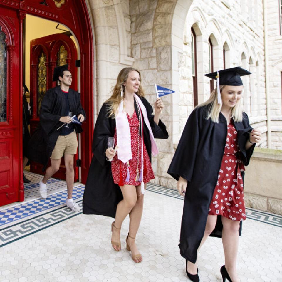 students in camp and gowns walking out of the main building red doors