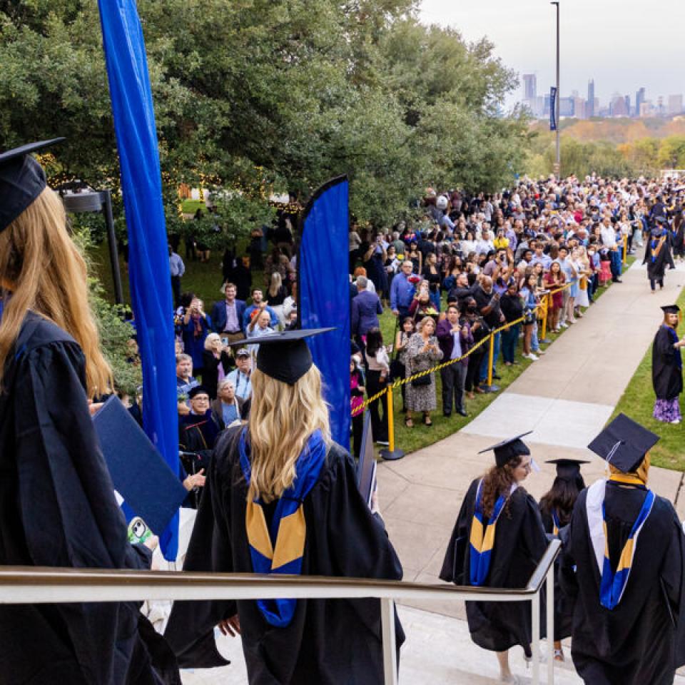 students in camp and gowns walking out of the red doors with the Austin skyline in view