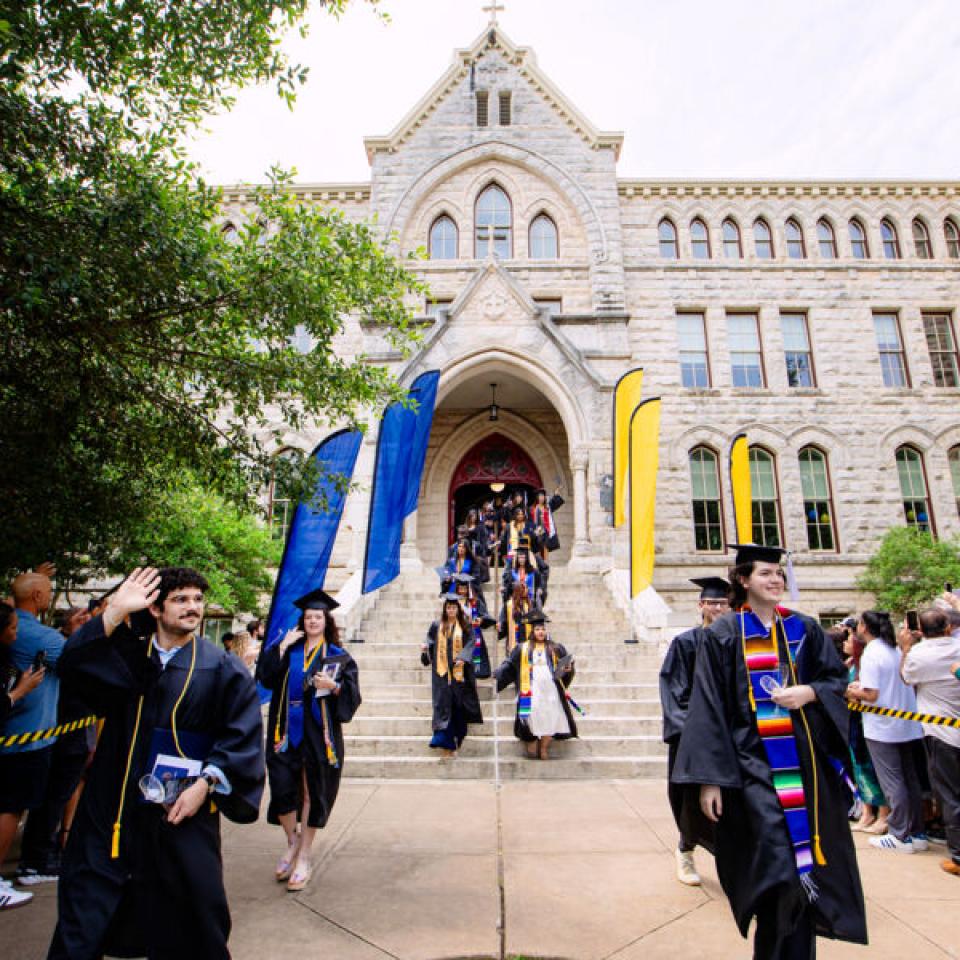 The image captures a graduation ceremony taking place outside a historic, stone-built academic building. Graduates in caps and gowns are walking down the steps, smiling and waving, with some wearing colorful stoles. The pathway is lined with cheering family and friends, separated by caution tape. Colorful banners are displayed along the steps, adding to the celebratory atmosphere. The scene is lively, filled with joy and accomplishment.