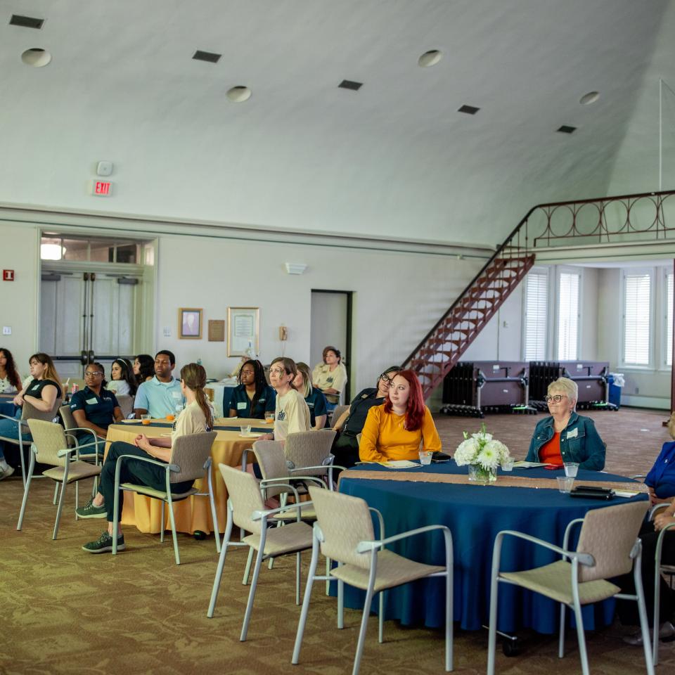 A group of people is seated at round tables covered with blue and yellow tablecloths in the Maloney Room. 