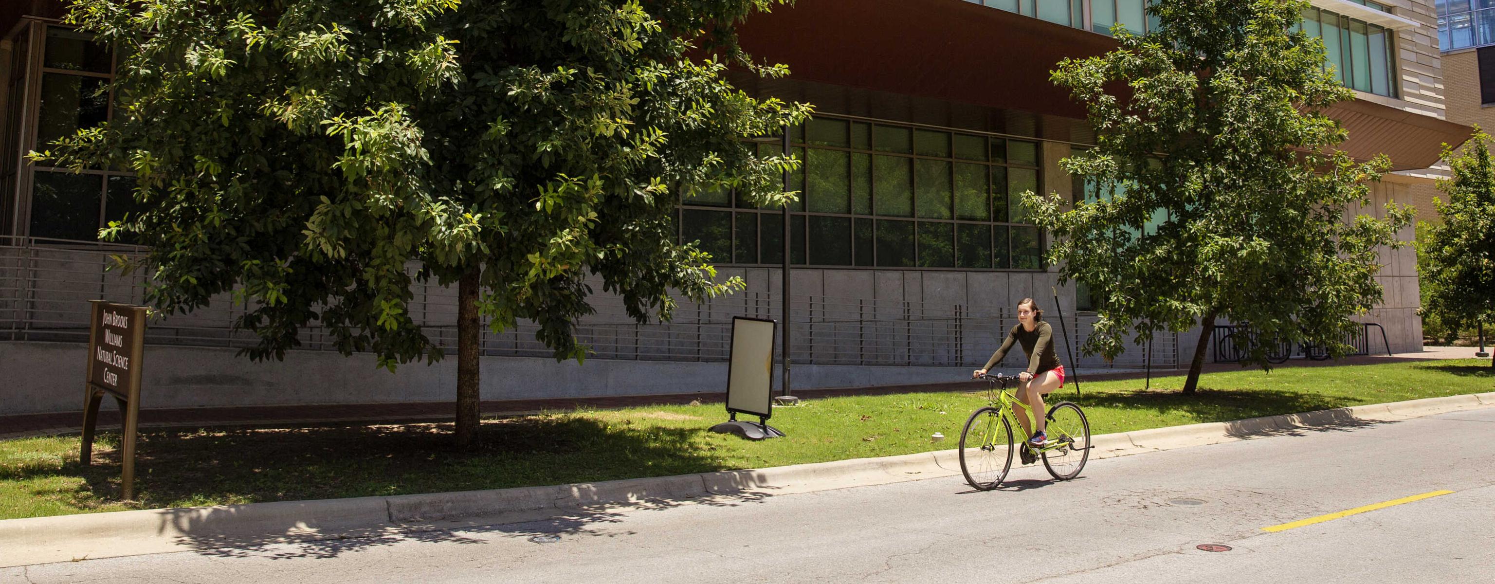 A student rides their bike on the street and is framed between two trees that are planted in front of John Brooks Williams Natural Sciences Center - South.