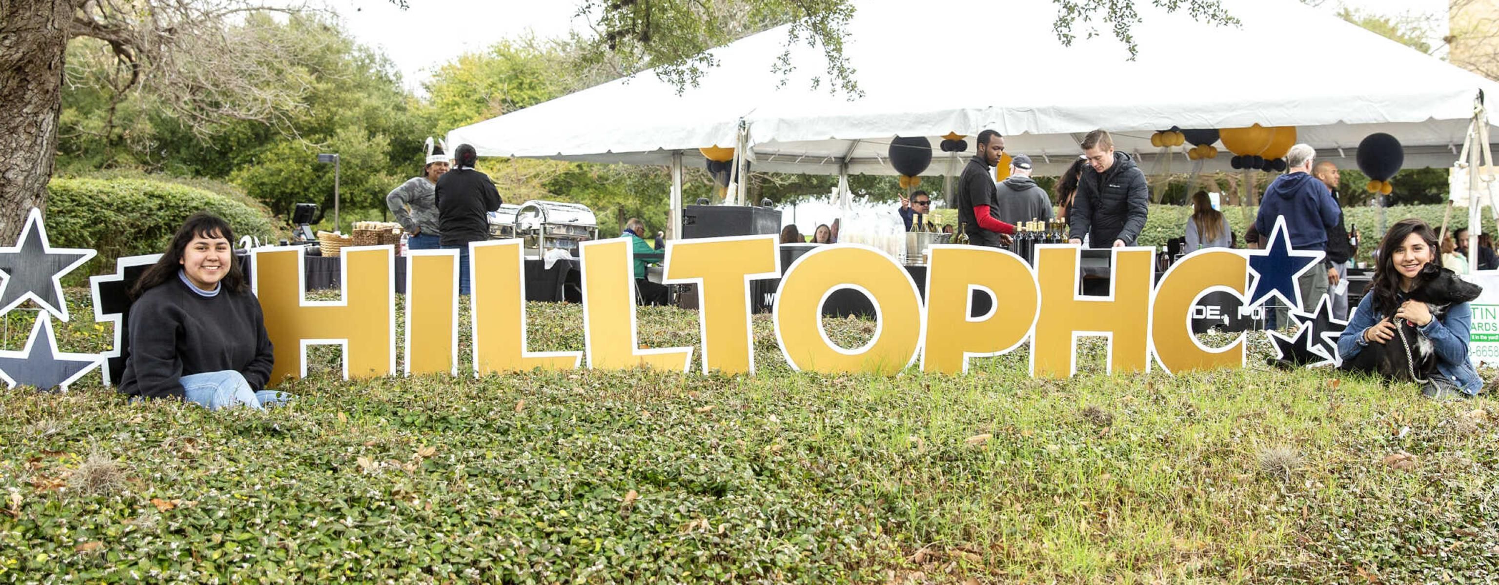 The image shows an outdoor event with a large sign spelling "HILLTOPHC" in bold, yellow letters. Two women are sitting on the grass, one on each side of the sign, smiling at the camera. One woman on the left has long dark hair, and the woman on the right is holding a small black dog. In the background, there is a white tent with people gathered around tables, indicating a festive atmosphere. The scene is set in a park-like area with trees and greenery.