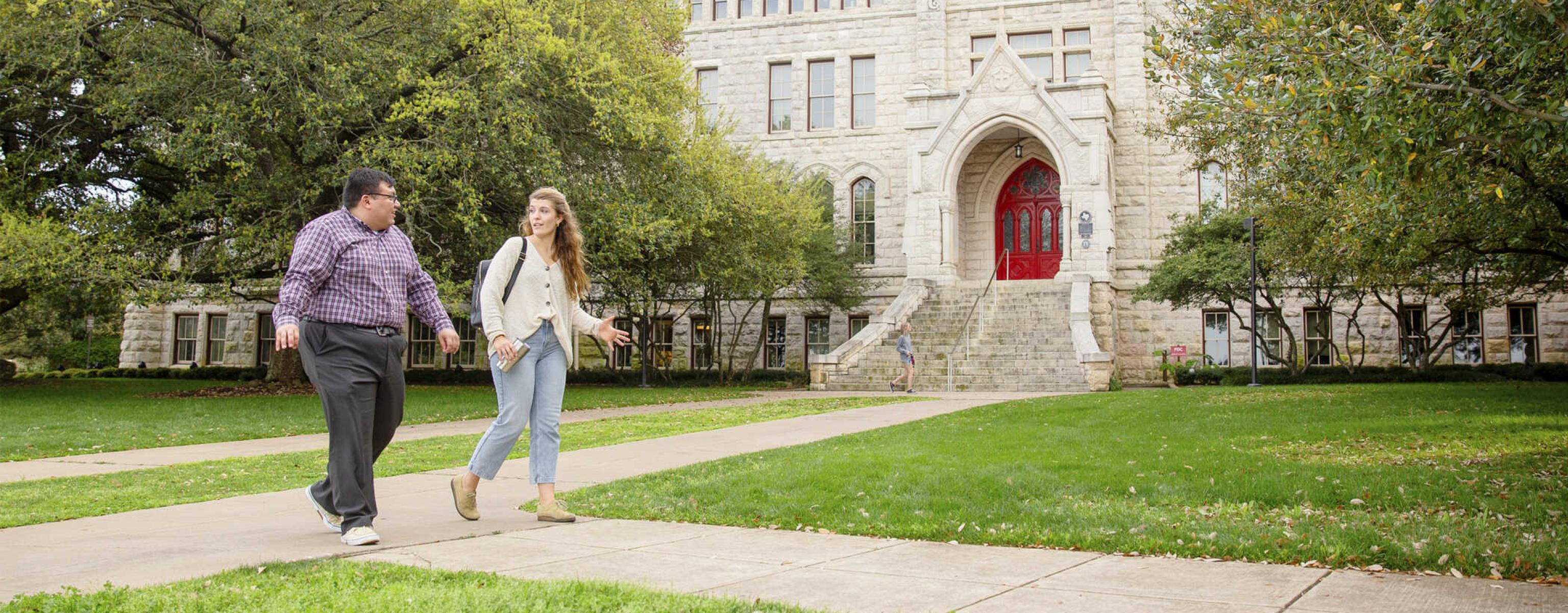 Two graduate students walk on a path in front of Main Building and oak trees.