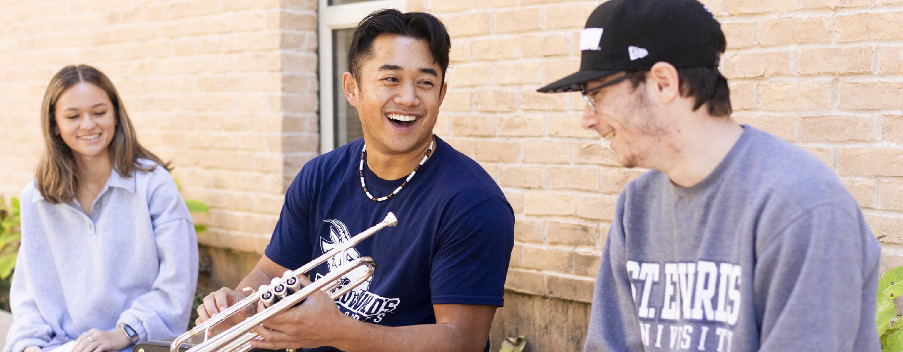 Three students sit and chat together outside of Trustee Hall. The student in the middle holds a trumpet.