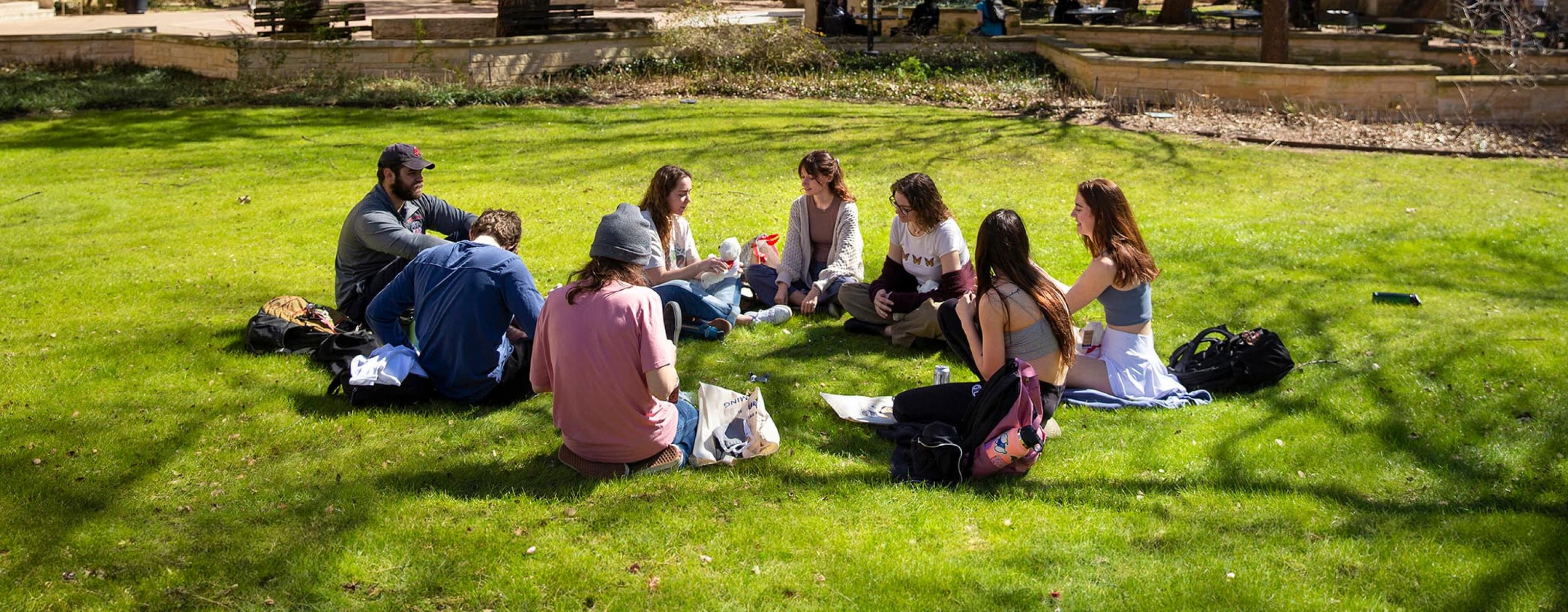 A group of students sitting on the grass in what appears to be a campus setting, with large trees providing shade and buildings in the background. The individuals are arranged in a circle, some with their backs to the camera, and appear to be engaged in a group activity or discussion. The scene is set on a sunny day with clear skies.