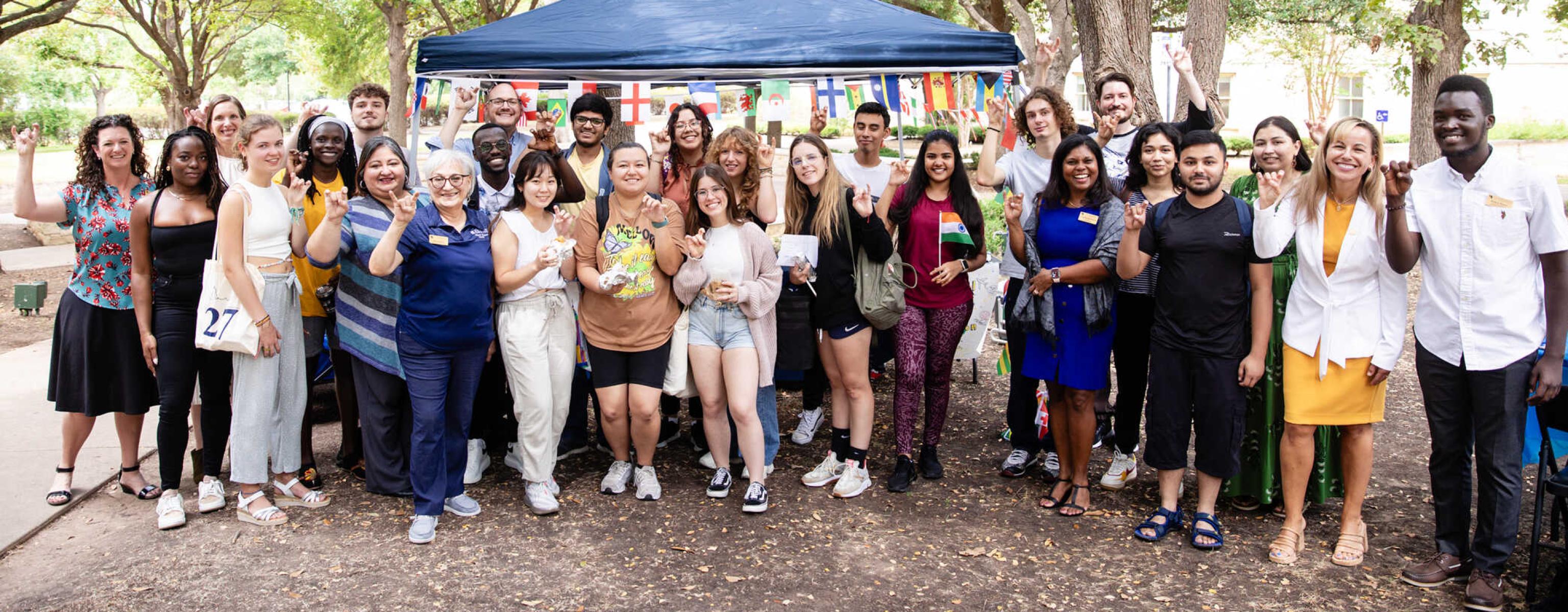 The image shows a diverse group of people gathered outdoors, smiling in front of a blue tent decorated with international flags. The setting is a park with trees, creating a relaxed atmosphere. Many participants are making the "Toppers Up" gesture, indicating a St. Edward's University event, likely for international students. The scene exudes unity, cultural diversity, and a celebratory, inclusive community spirit.