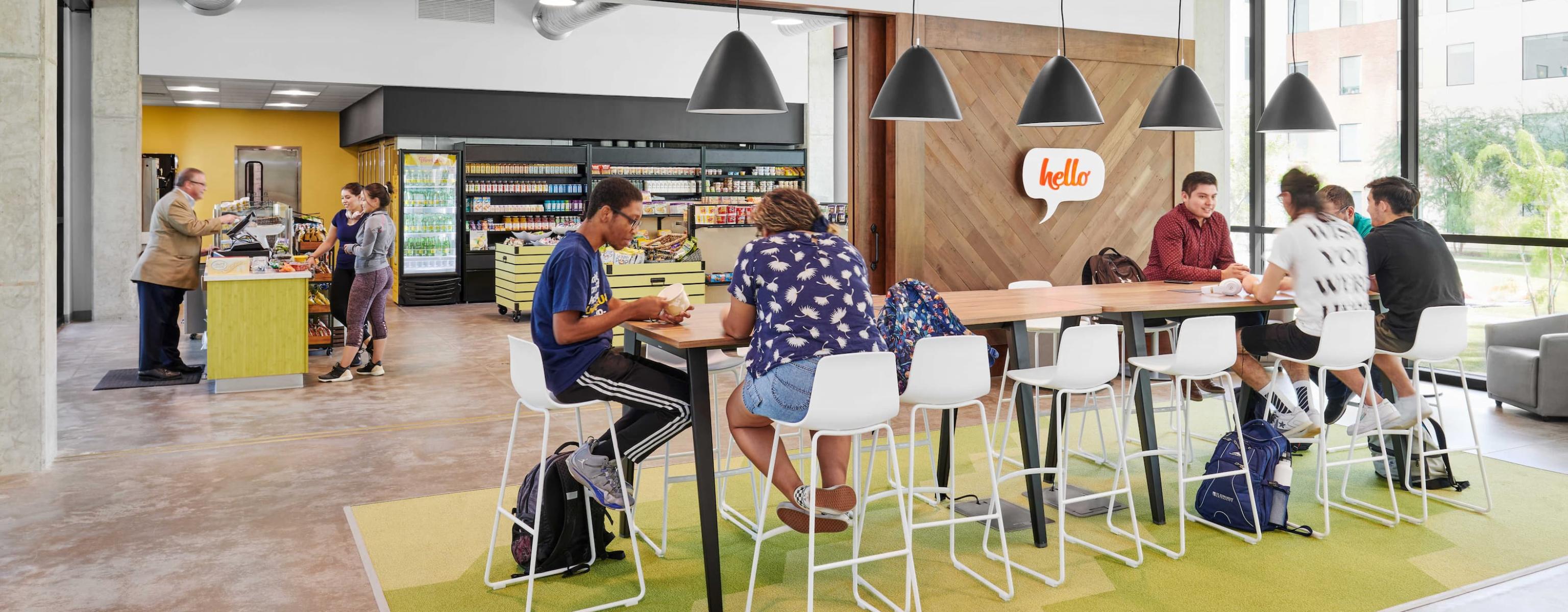 The image shows a modern café/lounge with a large communal table and pendant lights. People are seated, studying, or socializing. To the left, there’s a convenience store with drinks and snacks. The space has an industrial design with exposed ductwork, concrete floors, and large windows. A welcoming “hello” sign is on the wooden accent wall.