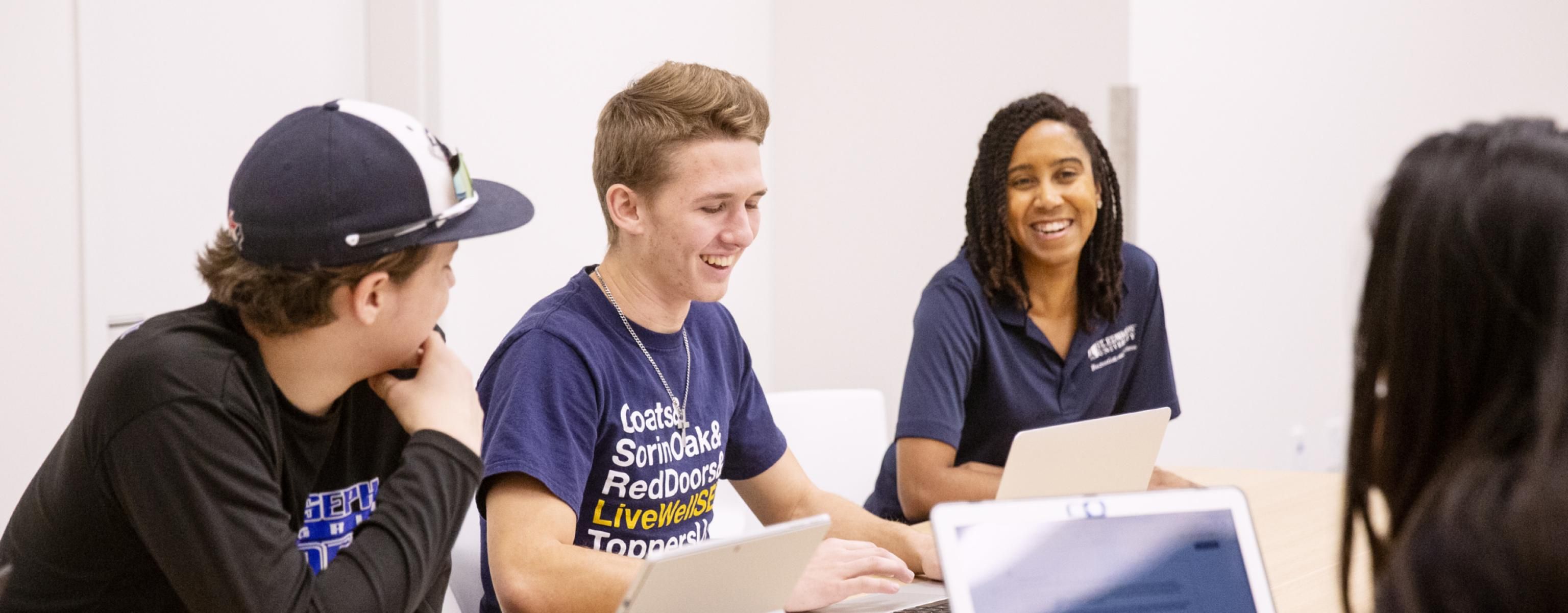 Students and a staff member smile during a meeting as they sit at a meeting table with laptops and tablets.