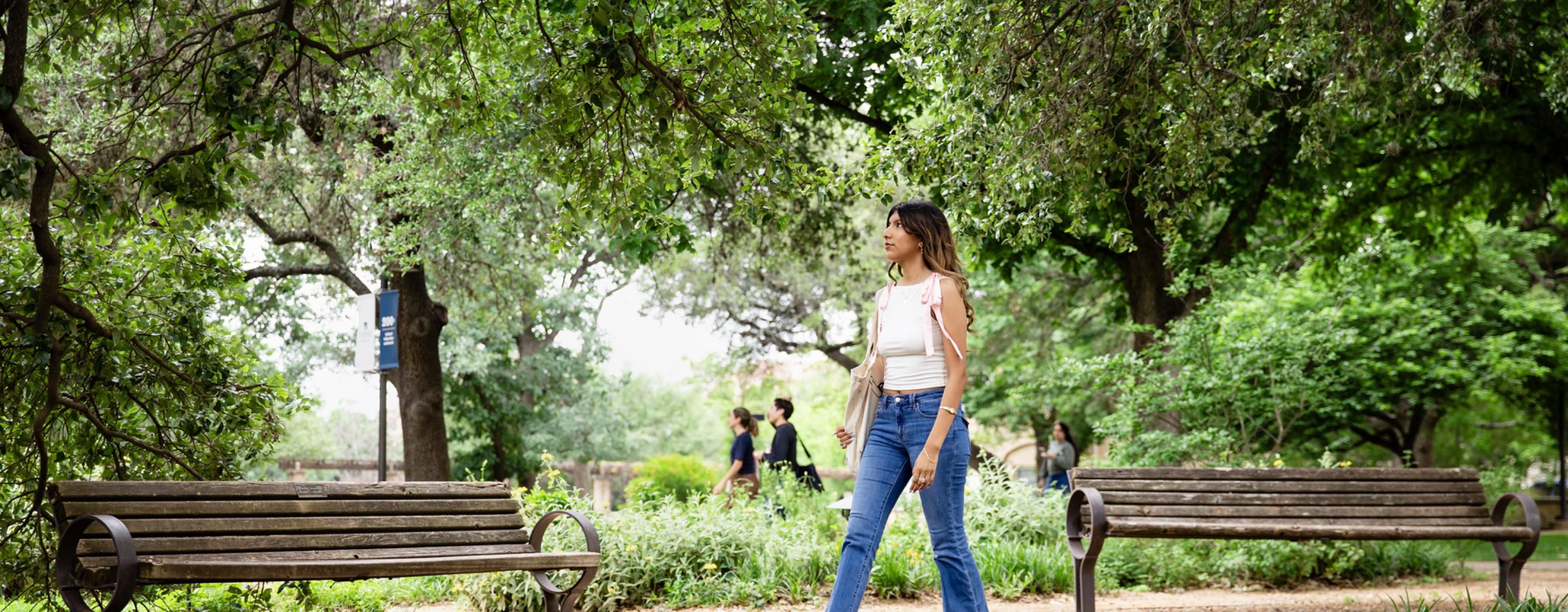 A student wearing a white shirt with pink ribbons on the shoulder and blue jeans walks on a stone gravel path, framed by benches and trees.