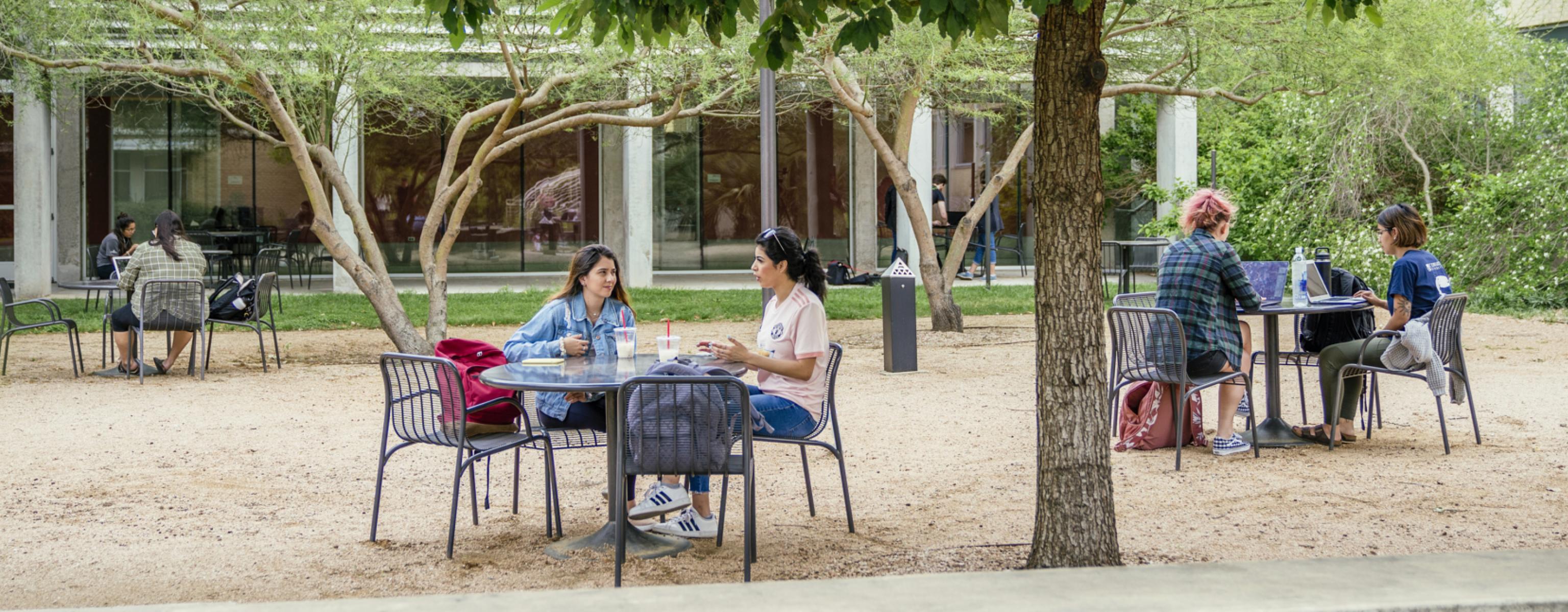 Students sit at tables in the Equity Hall courtyard, with backpacks and laptops, hanging out with friends and studying.