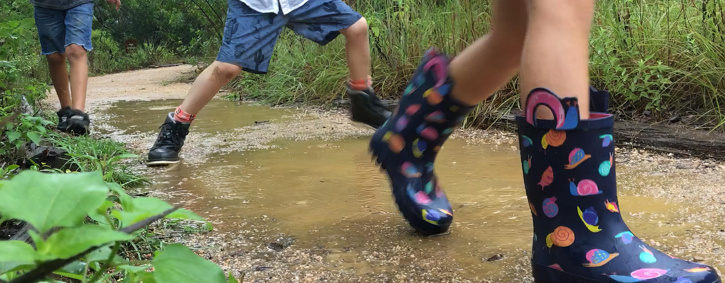 The image depicts three children on a dirt path. The foreground child wears colorful boots and steps into a puddle, creating ripples. Another child, wearing dark shoes and shorts, is mid-step. In the background, a third child waits to jump. Green plants line the path, suggesting an outdoor setting, possibly after rain.