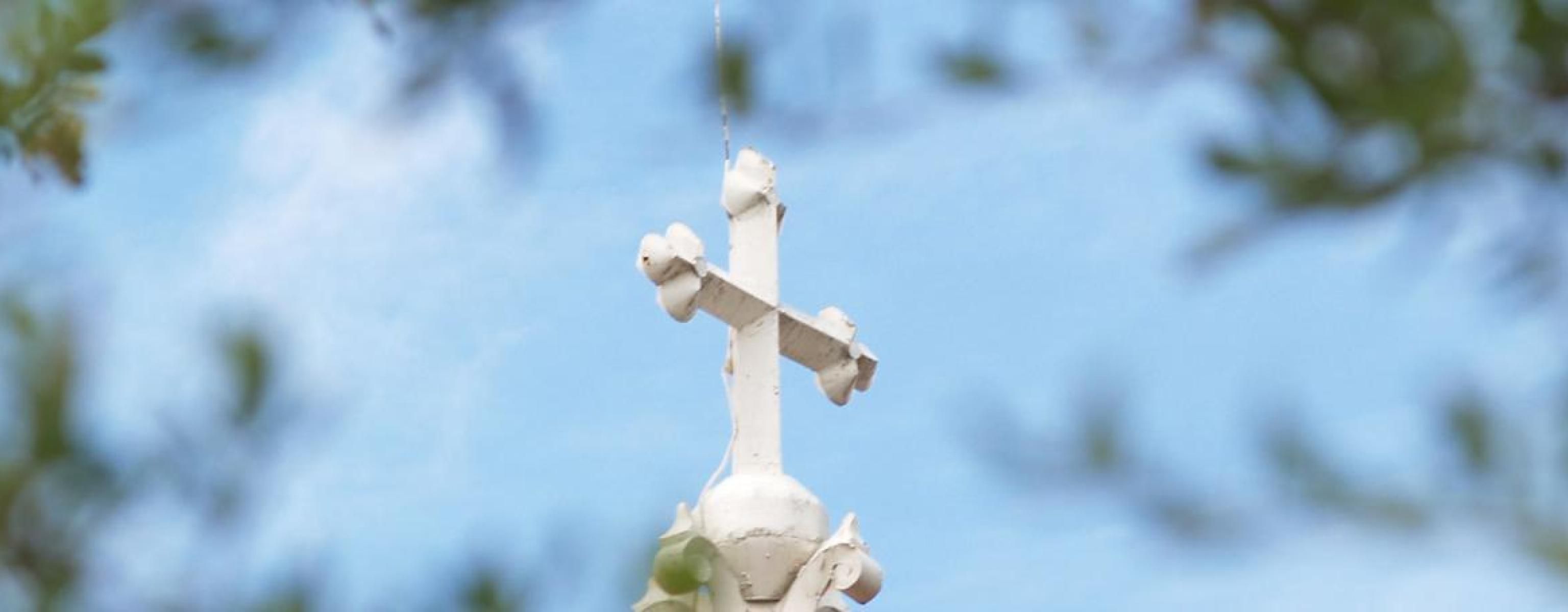 The image depicts a white cross atop a building, set against a blue sky. The cross has decorative elements and is viewed through a soft focus of green leaves, creating a frame around it. The scene conveys a peaceful and serene atmosphere, highlighting the cross as a central symbol of faith and spirituality.