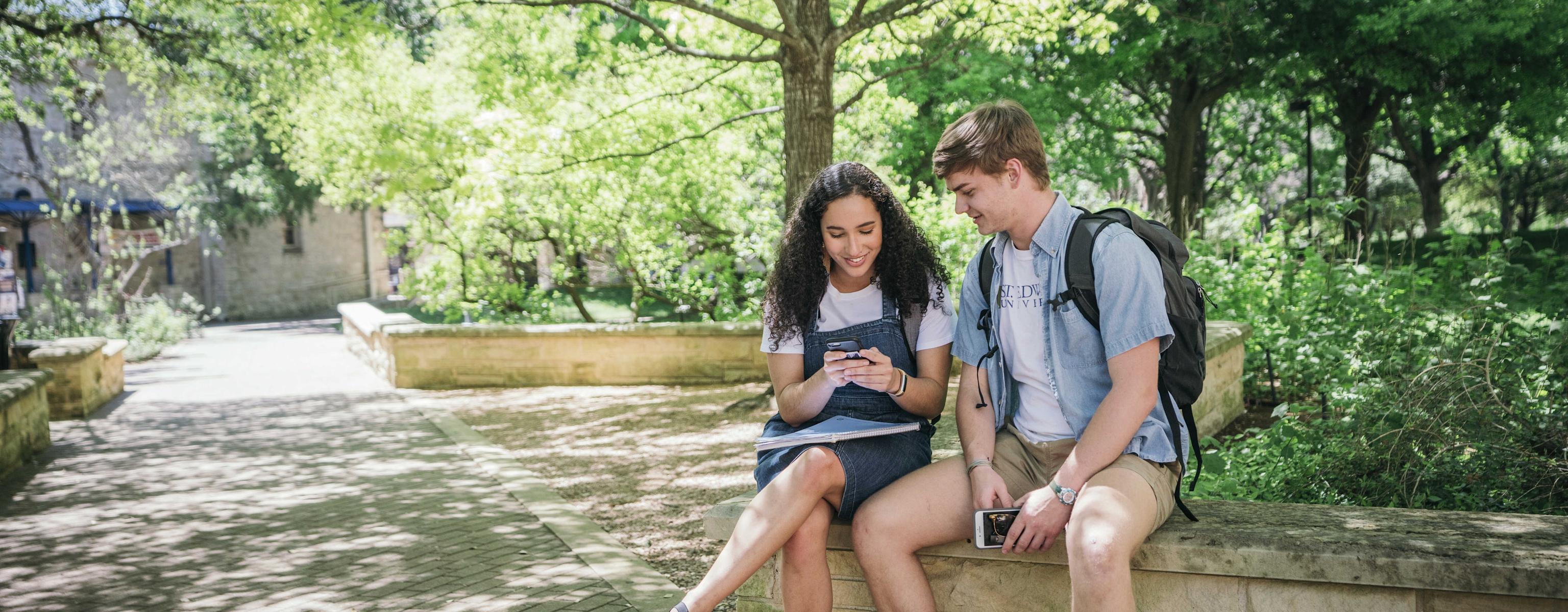 two students sit on a stone bench by a tree-lined walkway and look at a phone.