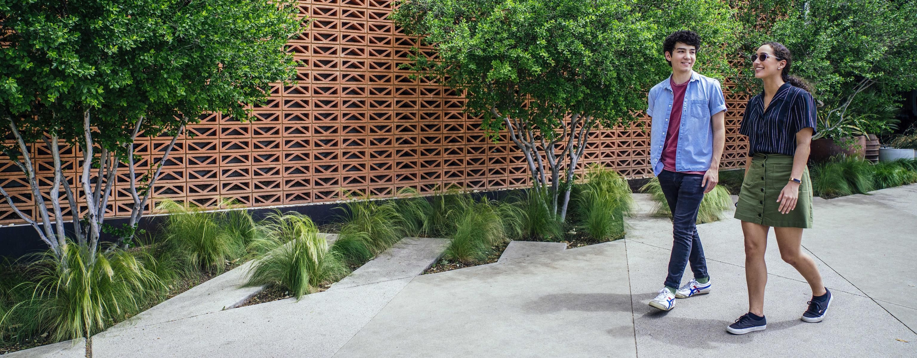 Two students walk by a earth-toned patterned stone wall with trees in front of it on South Congress.