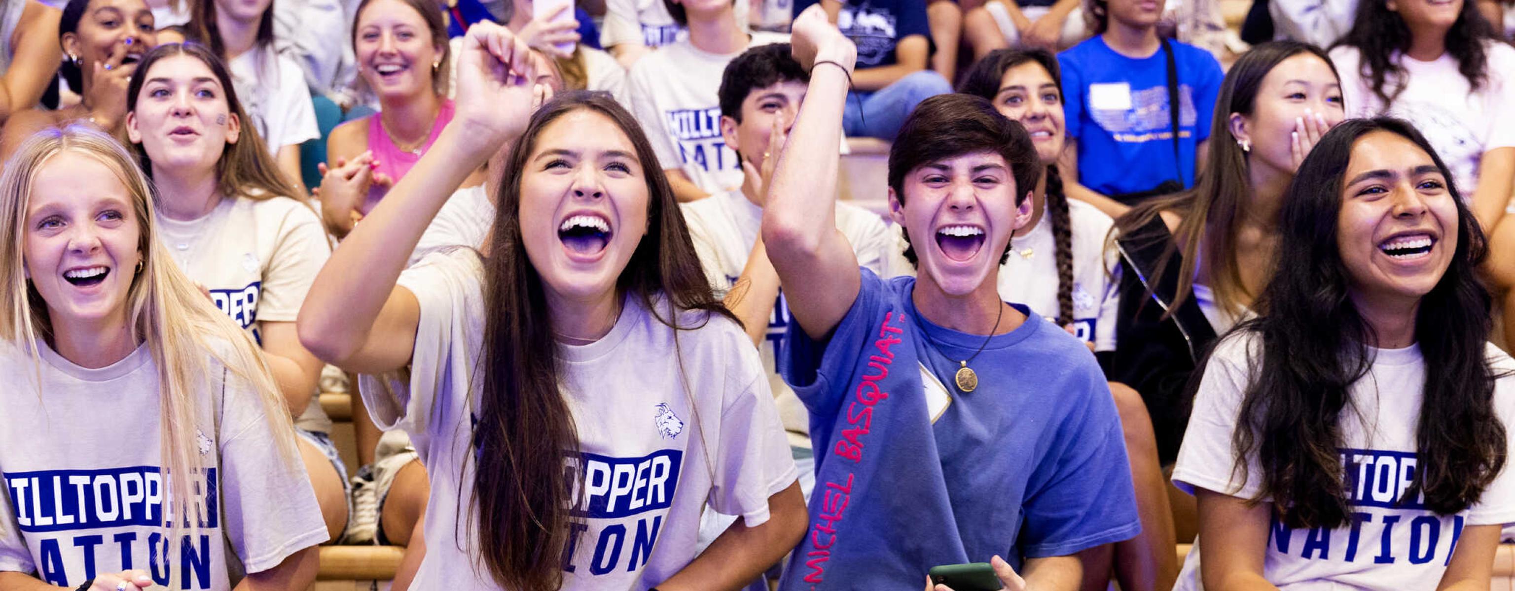 A group of students sit in the bleachers, many of whom are wearing Hilltopper Nation shirts, screaming and cheering during a pep rally.