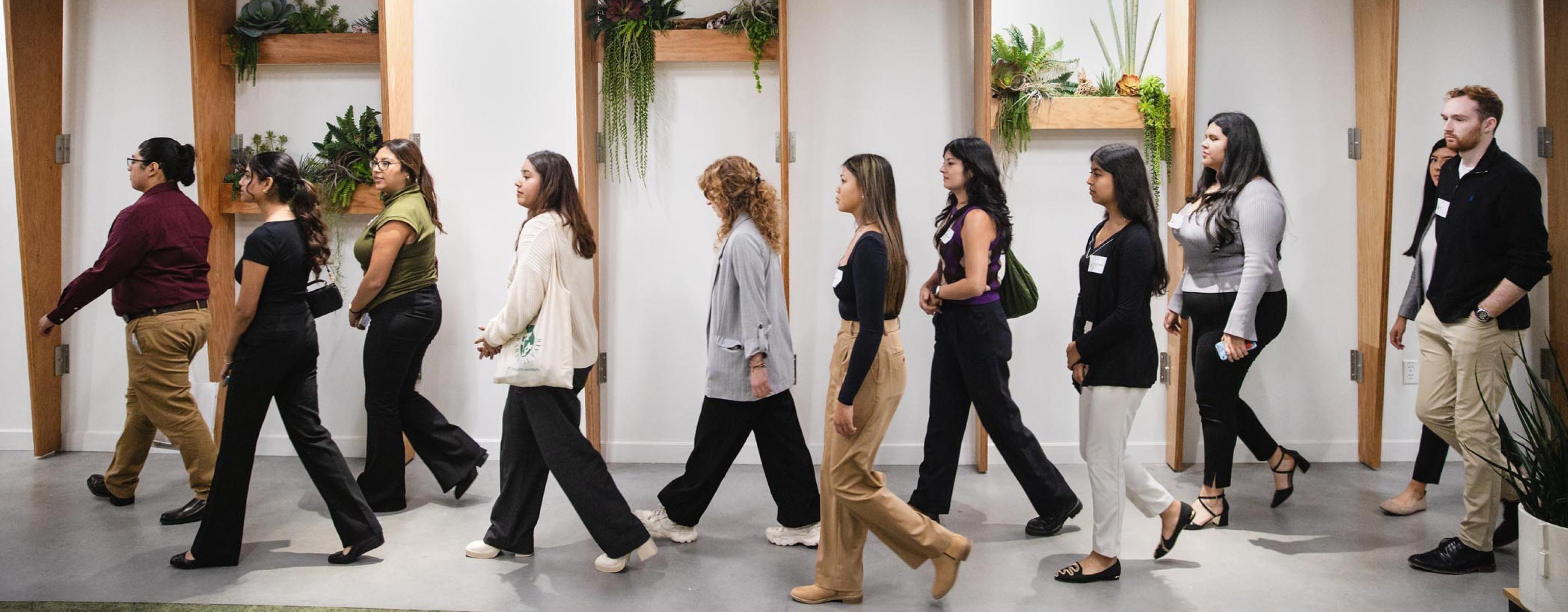 The image shows a group of professionally dressed individuals walking in a single file line indoors. The setting appears to be modern, with wooden beams and minimalistic design elements. The background features decorative shelves with plants, adding a touch of greenery to the space. The group consists of men and women, likely participating in a tour, workshop, or professional development event. The atmosphere is organized and focused.